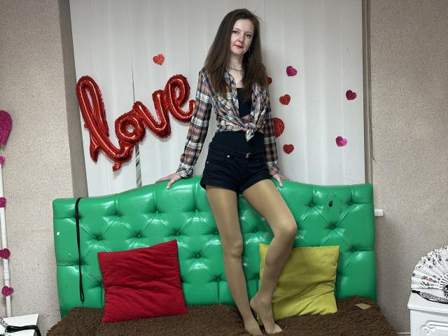 MeganPirs with long brown hair, plaid shirt tied at the front, black shorts, and tan stockings, standing on a green tufted couch with red and yellow pillows.
