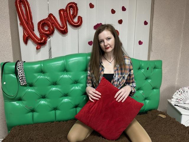 MeganPirs, a fair-skinned woman with long brown hair and red lipstick, sits on a green tufted leather couch. She wears a plaid shirt over a black top, and beige tights. She holds a red velvet pillow with both hands. A "Love" balloon and red rose petals are in the background. A black-and-white patterned bag and a white decorative box are on the right. The brown textured rug adds contrast to the green couch.