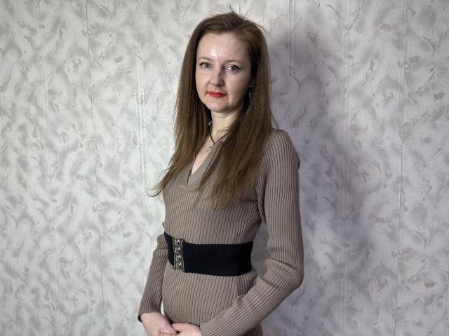 MeganPirs with long brown hair, red lipstick, wearing a ribbed brown dress with a black belt, standing, hands in front, looking at camera.