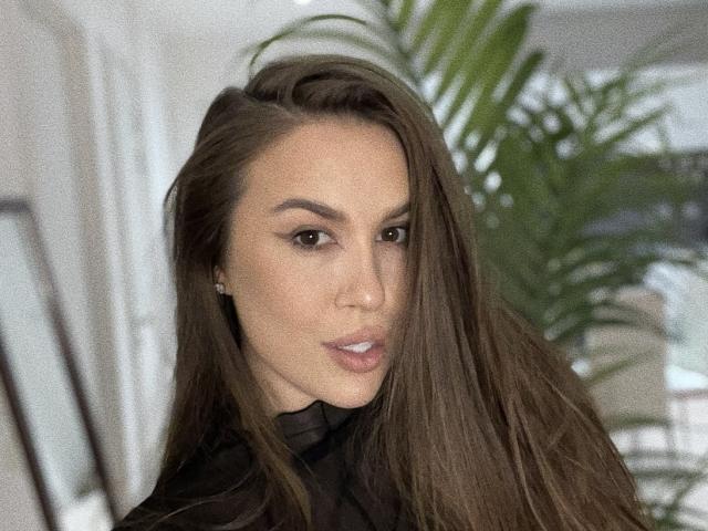 MirraBellle with long brown hair, subtle makeup, and a serious expression, wearing a black top, showing her nose and slightly parted lips, with green plant in the background.