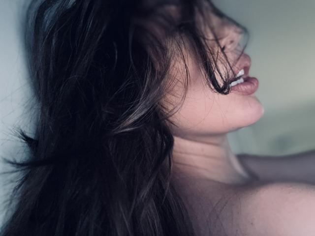 MirraBellle's face is partially obscured by her long, dark, messy hair. Her eyes are closed, and her lips are slightly parted, showing her upper teeth. The hair covers her right eye and falls over her forehead. The light source is soft, creating a gentle shadow on her left cheek. Her skin is pale, and the composition focuses on her upper face and neck. The image has a moody, intimate feel.