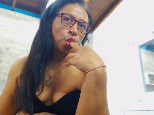 SoffieMature with long black hair, red glasses, and black top, sucking her finger with her red-lipsticked mouth, showing her cleavage and necklace. Her ass is slightly visible.