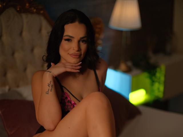 NayraZahra with dark wavy hair, wearing black and pink lace lingerie, sitting on a bed, hand on chin, tattoo on wrist, soft smile, dimly lit, warm ambiance.