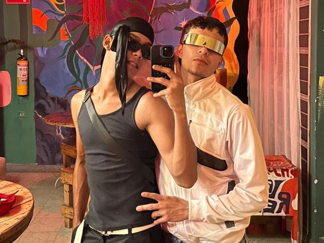 DanAndKamadeva with black mask, black tank, and black shorts, standing next to Kamadeva with white jacket, gold headband, and black strap, taking selfie. Kamadeva's hand on DanAndKamadeva's ass.