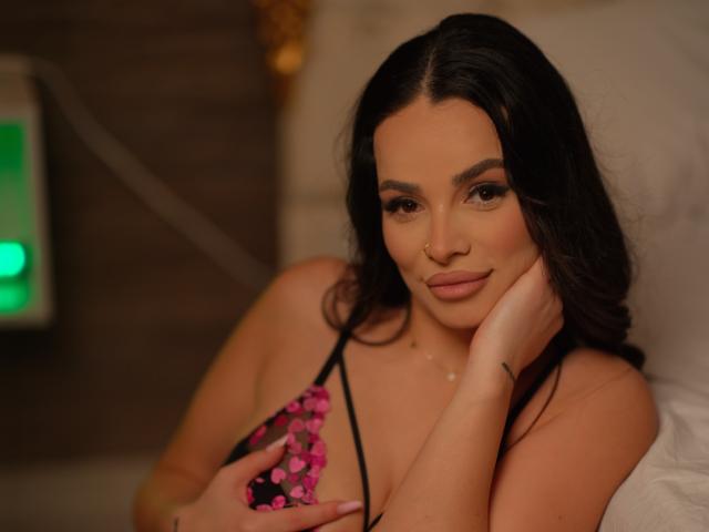 NayraZahra with dark hair, brown eyes, and pink floral bikini top, gently touching her cheek, smiling subtly, nose ring visible, in a warm, softly-lit setting.