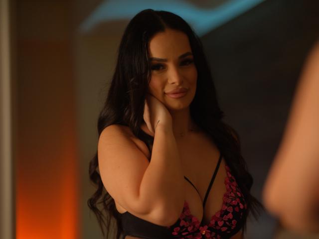 NayraZahra with long black hair, smiling, touching her cheek, wearing a black and pink floral bra that accentuates her big boobs. Soft, warm lighting. Brazers watermark in bottom right.
