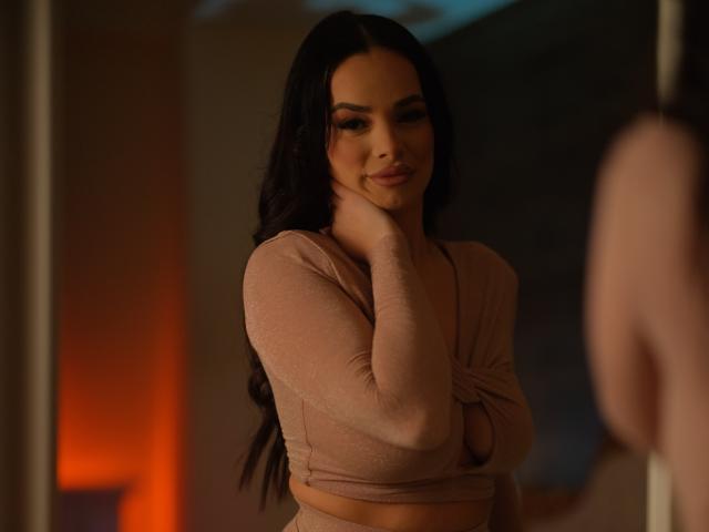 NayraZahra with long black hair, wearing a pink long-sleeve top, touching her chin with a slight smile, her tits partially visible, in a dimly lit room.