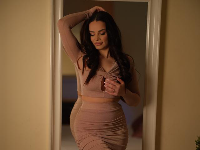 NayraZahra with long dark hair, wearing a pink, long-sleeve crop top and ruched skirt, standing in a doorway, gently touching her tits, smiling softly.