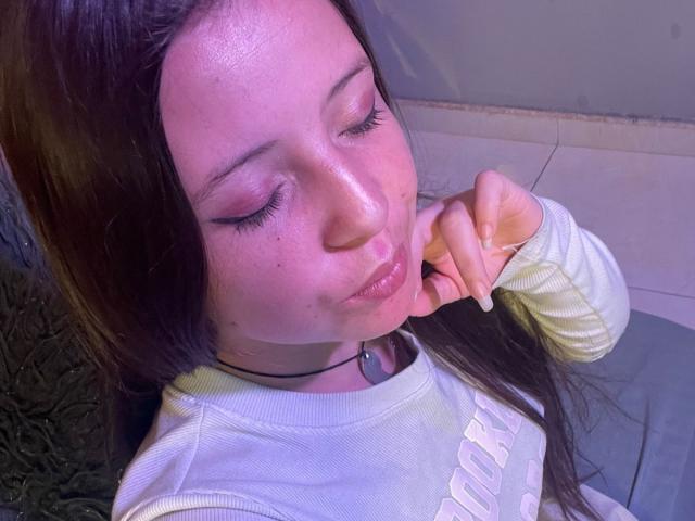 A close-up of GraceRedMiller with fair skin, long brown hair, and subtle freckles. She wears a light yellow ribbed sweater with white text. Her eyes are closed, and she pouts her lips slightly. She holds her right hand near her chin. Her eyelashes are long and dark. She has a black choker necklace with a small circular pendant. The lighting is pinkish, highlighting her smooth, shiny skin.