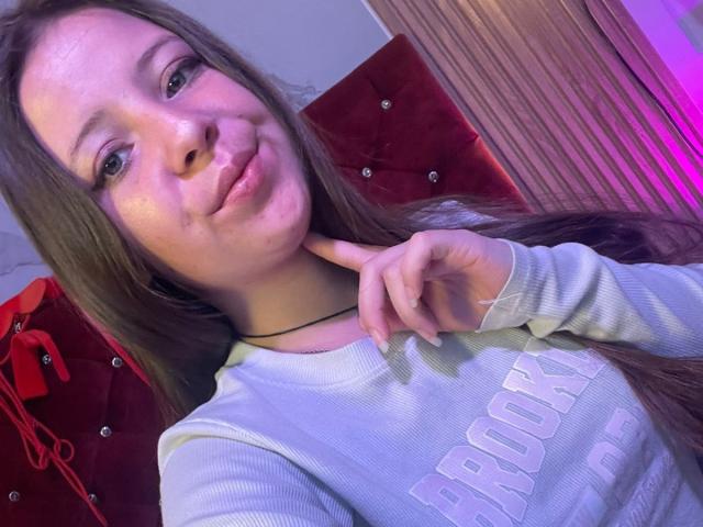 GraceRedMiller has long brown hair and fair skin, with subtle freckles on her cheeks. She wears a white ribbed shirt with "PROCK" in large letters. She's holding her chin with her right index finger, slightly smiling. The background includes a red, tufted headboard and pink-purple lighting. Her eyes are blue, and she has a black choker around her neck. The image is a close-up, focusing on her upper body and face.