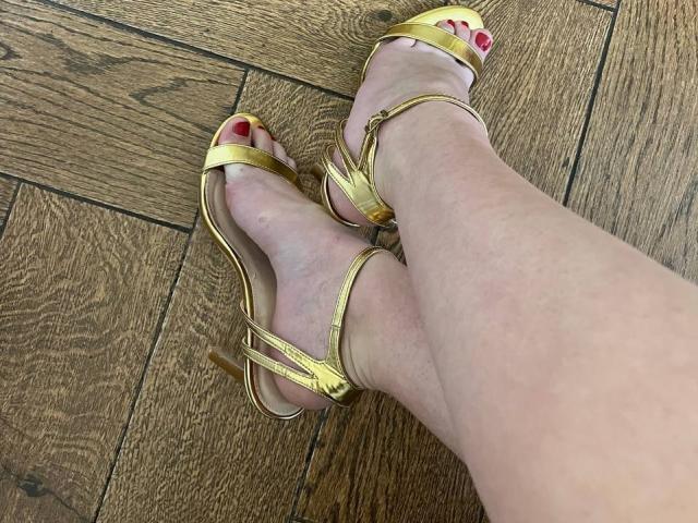 EolantaMarina's feet in gold strappy sandals with red toenails, slightly crossed, on wooden floor. Visible ankle and lower leg, pale skin.