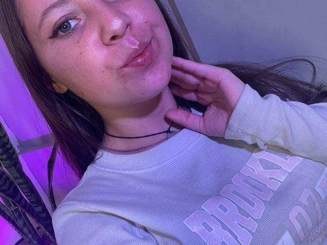 GraceRedMiller has shiny pink lips, a slight pout, and is wearing a white ribbed shirt with a text print, touching her neck with a hand, purple light behind.