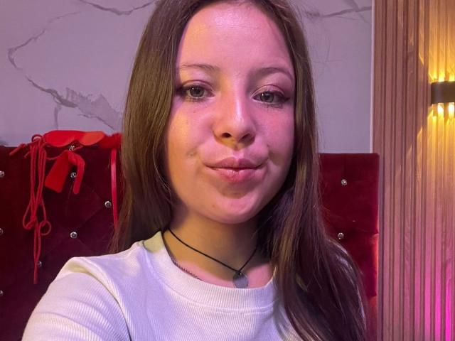 GraceRedMiller, con labios carnosos, vestida con una camisa blanca, cabello castaño y un collar negro, sonriendo ligeramente, en un fondo rojo de terciopelo con una cinta roja y una lámpara de pared..