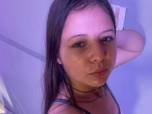 Close-up of GraceRedMiller, a young woman with pale skin, dark brown wet hair, and small freckles on her face. She wears a black choker and a thin black strap. Her right arm is raised, hand behind her head. She has dark, slightly downcast eyes, and her lips are slightly pursed. The image has a purple-blue tint. Her expression is neutral, slightly serious. The photograph is candid and unfiltered.