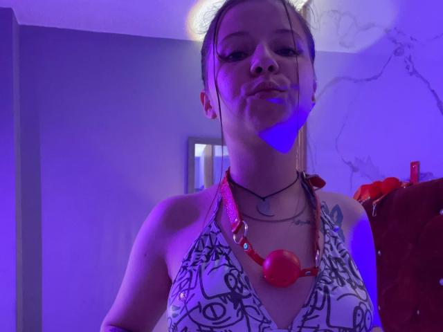 GraceRedMiller has a confident, slightly smug expression. She's wearing a white patterned halter top and a red necklace with a large red ball. Her hair is pulled back, and she has tattoos on her right shoulder. Blue and purple light casts a glow on her face. She's in a room with a framed picture and a red chair in the background. The light creates a dramatic, almost theatrical effect.