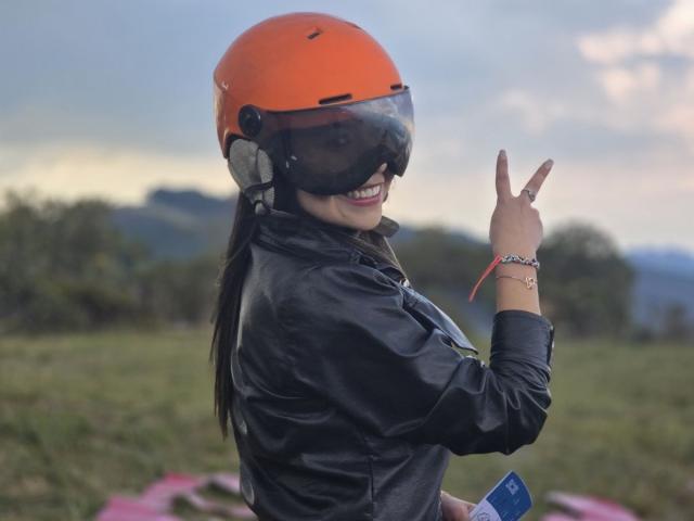 AshleyyPort con casco arancione, giacca di pelle nera e occhiali, sorridente, che fa il gesto della "rock on" con la mano, tenendo una carta blu, all'aperto, con erba verde..