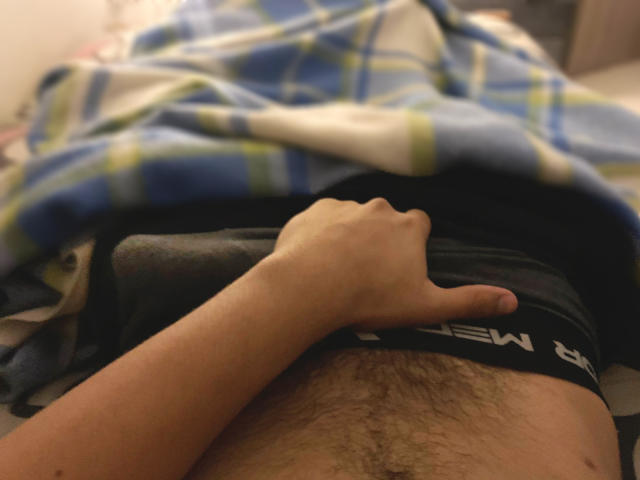 Close-up of AdoniBaskerville's lower torso, lying down. AdoniBaskerville's right hand, with a light skin tone, gives a thumbs-up over their black underwear, which has "NEW" text on the waistband. AdoniBaskerville's hairy abdomen is visible. A blue and green plaid blanket is partially covering AdoniBaskerville's body. The image is slightly blurry, with a warm, dim light.