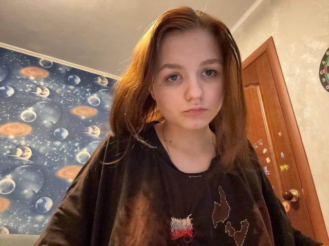 ClareBoulos has pale skin, brown hair, and wears a black tie-dye shirt with a small red and white pattern. She looks serious with a slightly parted mouth.