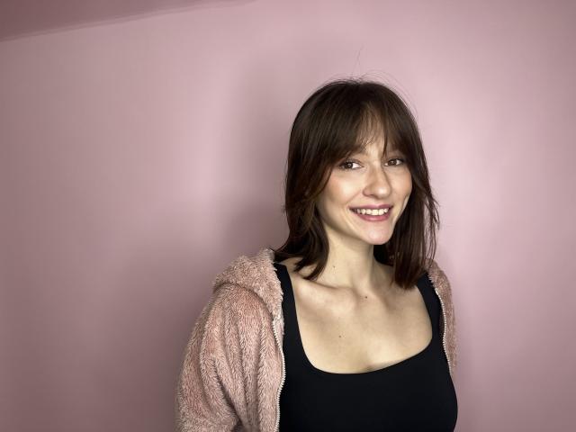 IlseJett ha capelli castano di media lunghezza, un viso sorridente e indossa un cardigan rosa e morbido sopra una canotta nera, che mette in mostra il suo seno piccolo e la sua figura snella..