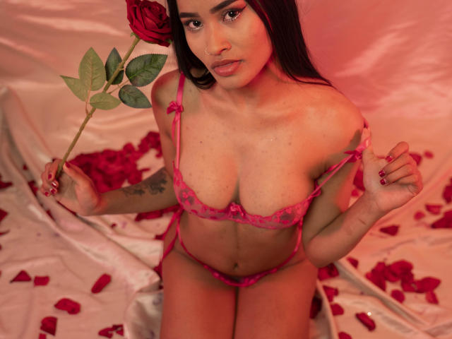 KarolPalacios with red rose, holding petals, wearing pink lace bra and panties, revealing her tits, sitting on white satin, her dark hair and red eyes contrasting the pink.