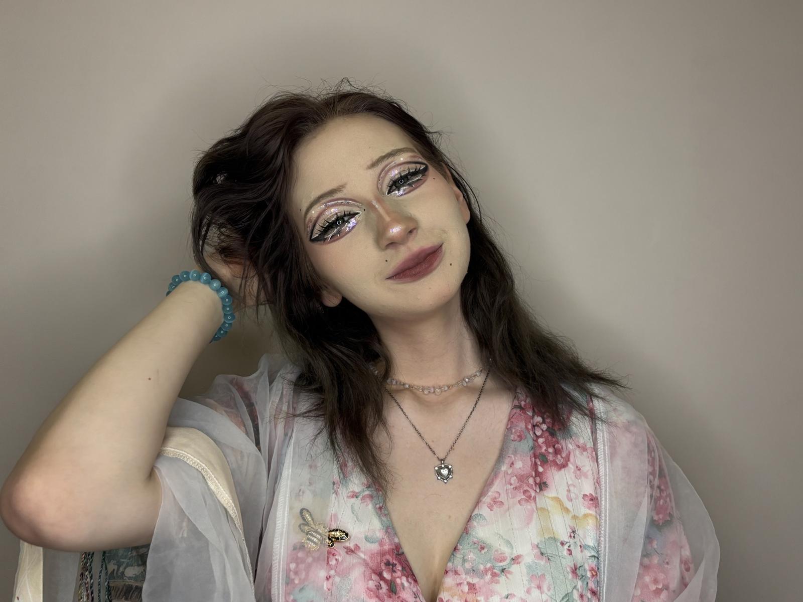 MiguelinaHoven has dark brown hair, wearing a floral pink and white top with a sheer white robe. She has blue eye makeup, a nose ring, and a blue bracelet.