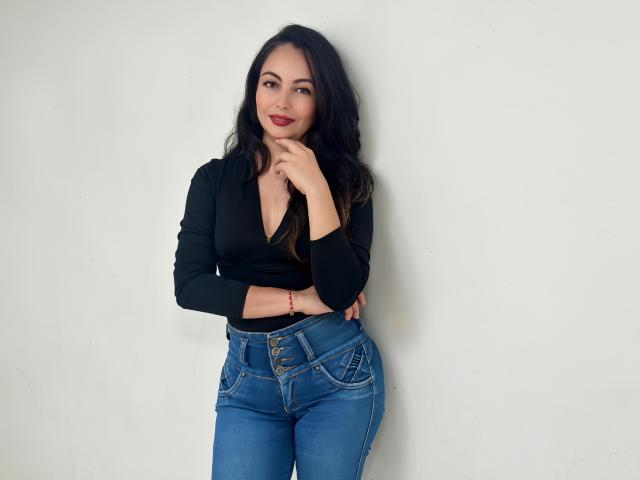 ToriBurns with long black hair, black V-neck top, and blue jeans, standing against a white wall, hand near chin, smiling, showing her tits and ass.