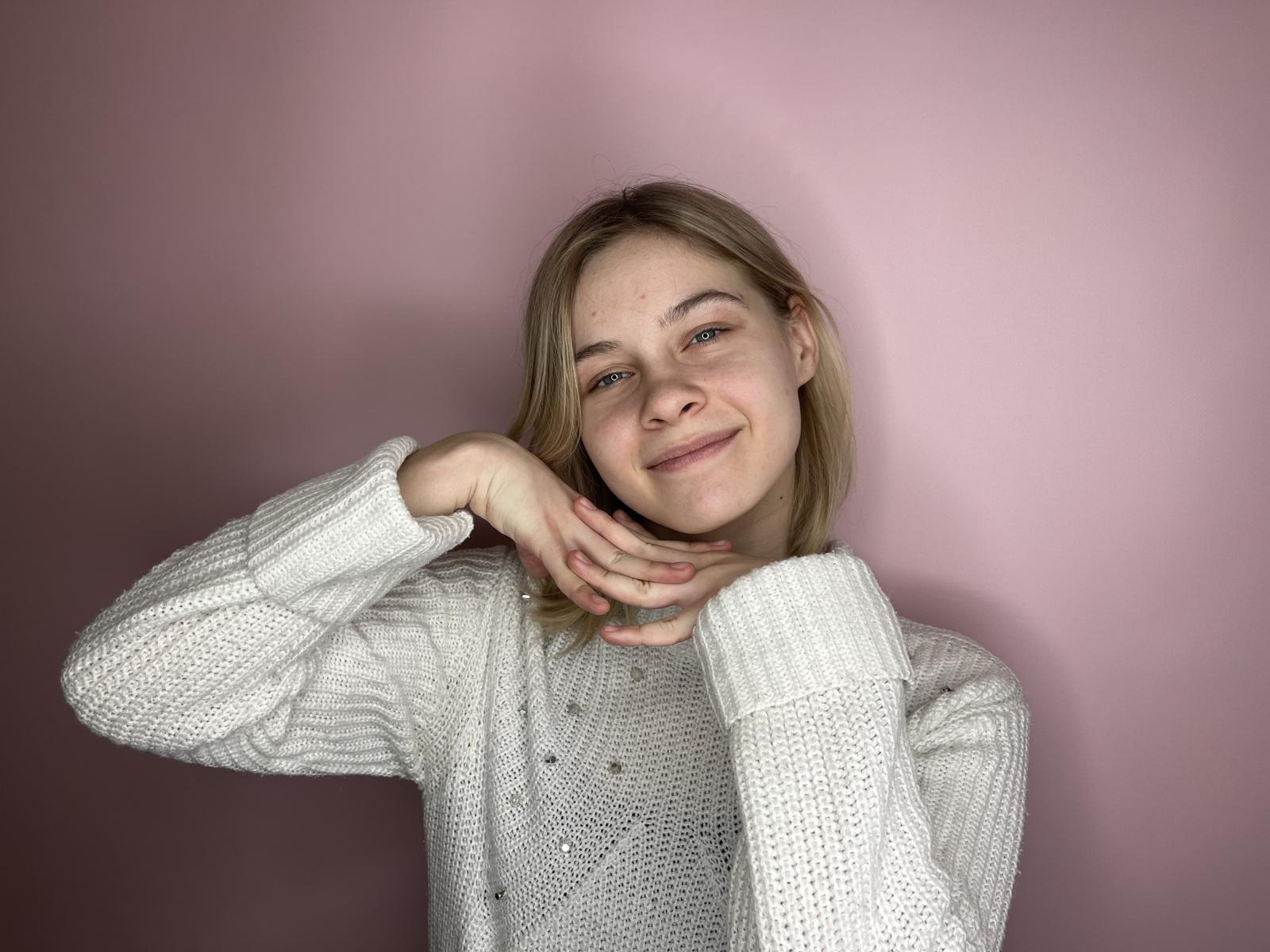 ShinPipes, con cabello rubio, sonriendo ligeramente, vestida con un suéter blanco de punto, tiene las manos tocando suavemente su barbilla y mejilla, fondo rosa..