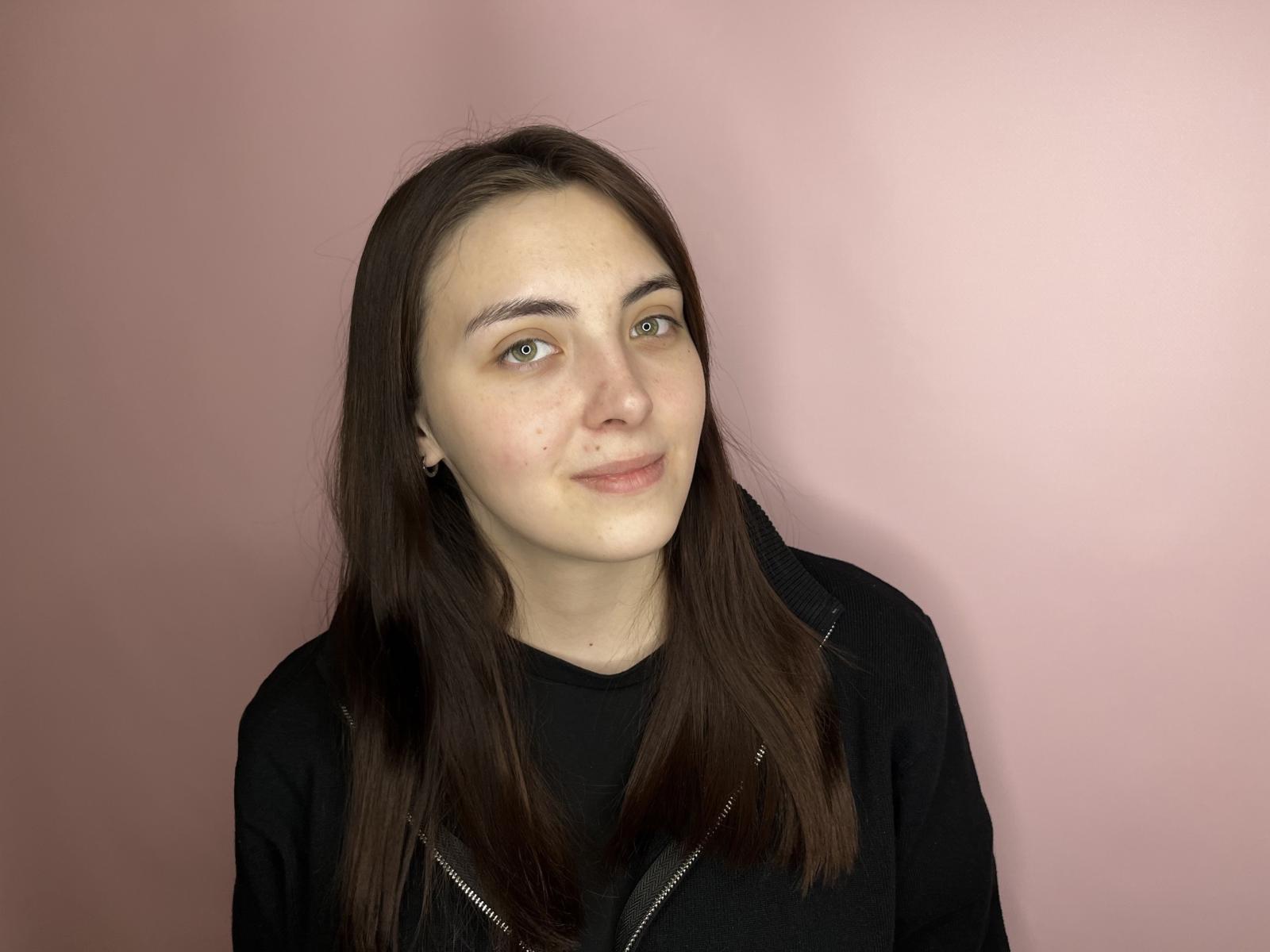 DaphineYuk tem pele clara, olhos verdes e cabelos castanhos longos. Ela está vestindo uma camisa preta com zíper, e está sorrindo levemente, com uma expressão relaxada..
