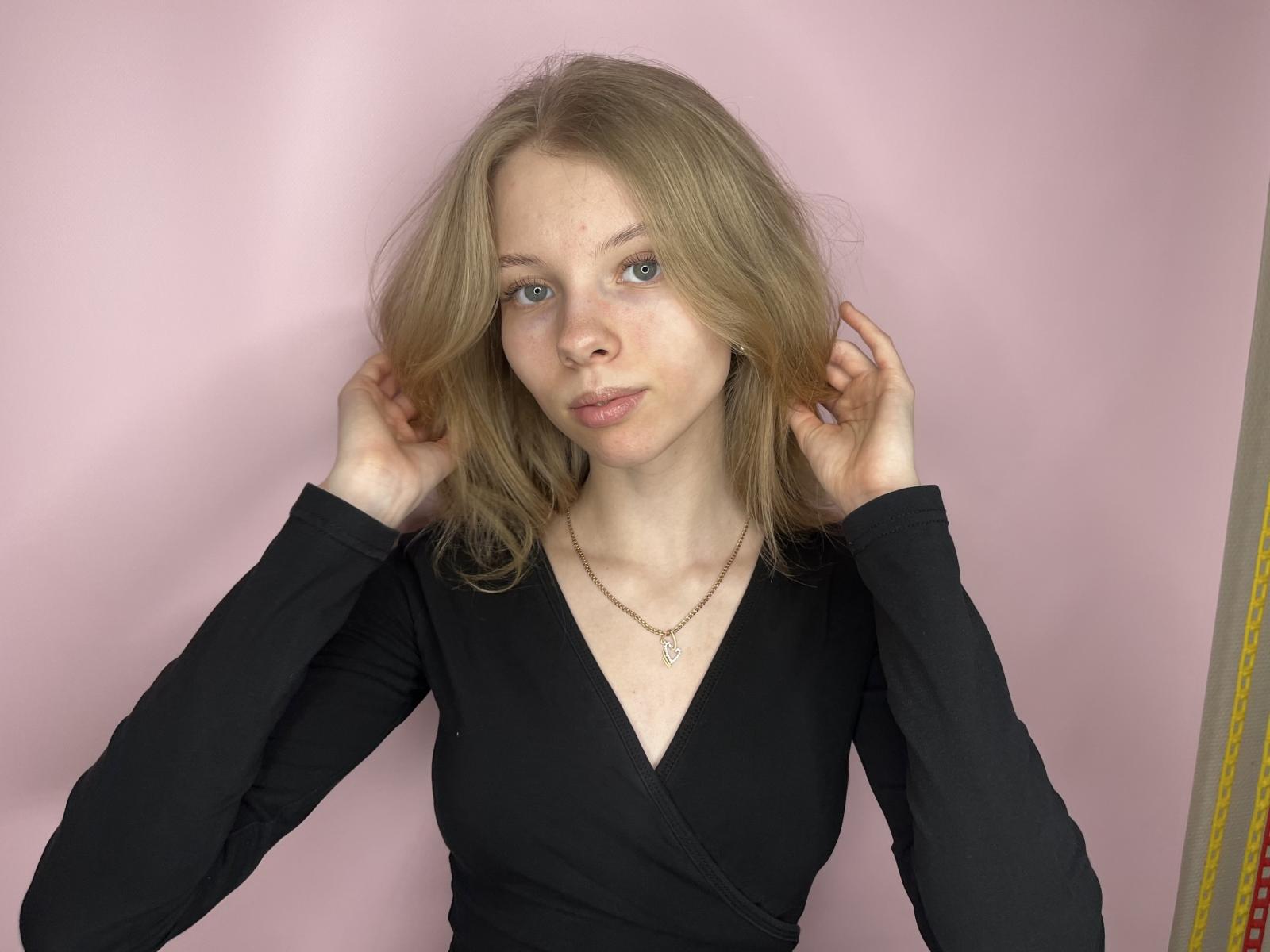 RiaMcabier has blonde hair, blue eyes, and fair skin. She's wearing a black long-sleeve top with a V-neck, hands in her hair, and a necklace.