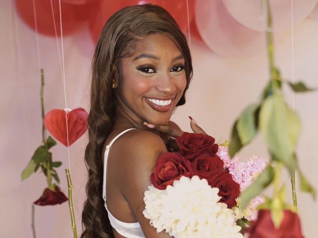 LiaTrainor with dark skin, long wavy brown hair, white dress, holding red and white roses, smiling with white teeth, red and pink balloons in blurred background.