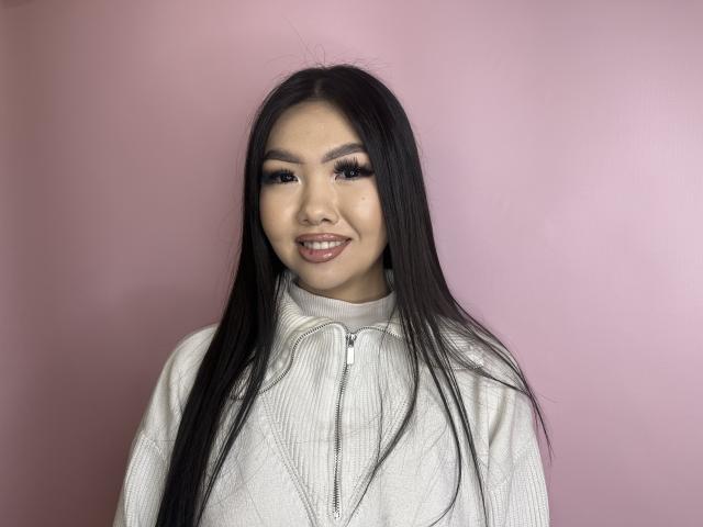 Giovanna Gossow heeft lang zwart haar, volle wenkbrauwen, dikke wimpers en ze glimlacht met haar glanzende, roze lippen. Ze draagt een witte sweater met rits, waardoor haar schattige gezicht te zien is..
