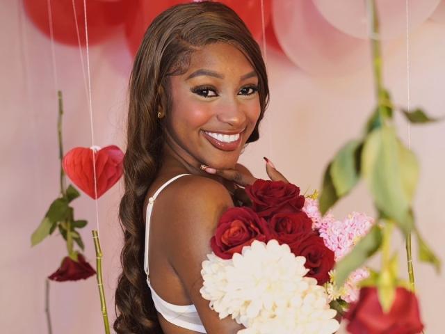LiaTrainor with dark brown skin, long wavy hair, white bra, holding red and white roses, smiling at camera, showing off her ass with a finger on her cheek.