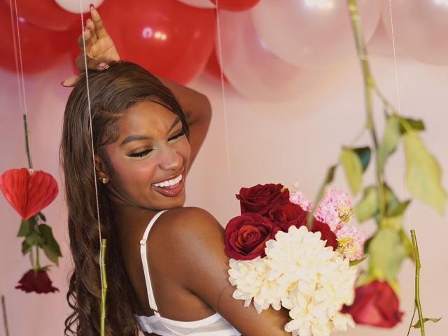 LiaTrainor with dark skin, long brown hair, smiling, holding roses and flowers, white strap dress, red and white balloons, one arm raised, looking back, showing her ass.