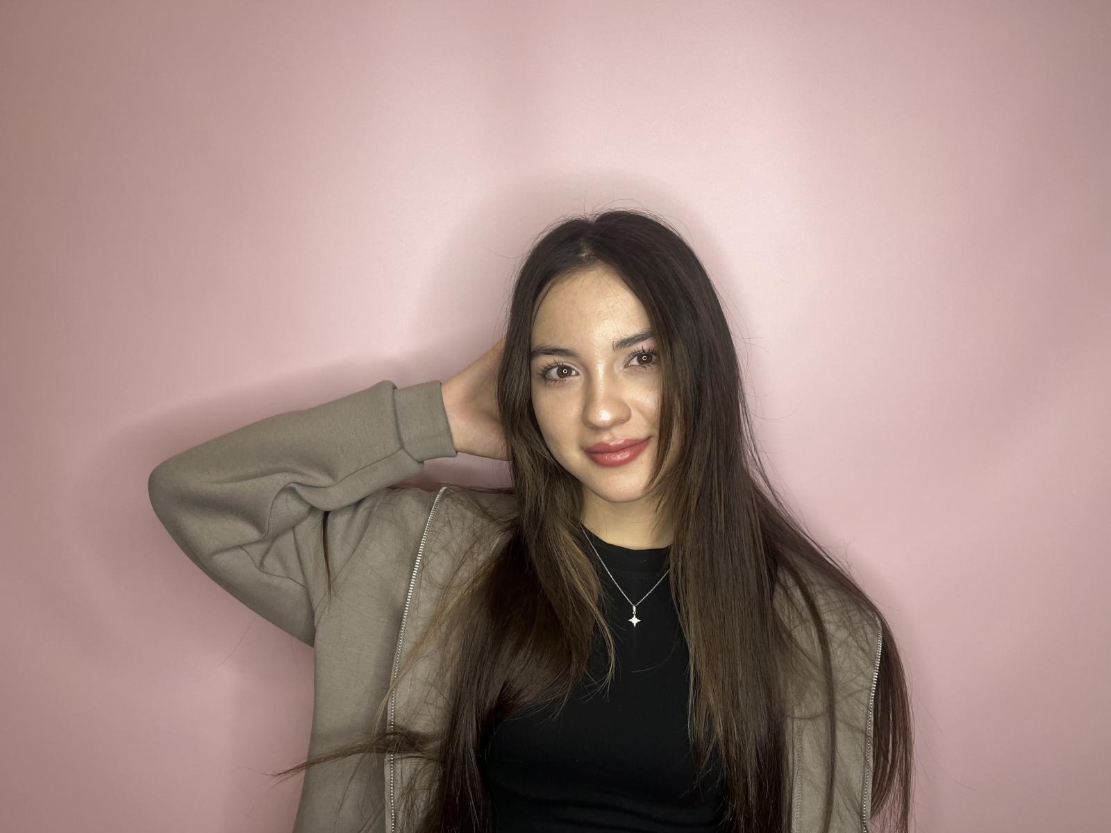 BrigetteMorelen with long brown hair, wearing a gray jacket over a black top, hand in hair, smiling, necklace with small star, pink wall, looking cute.
