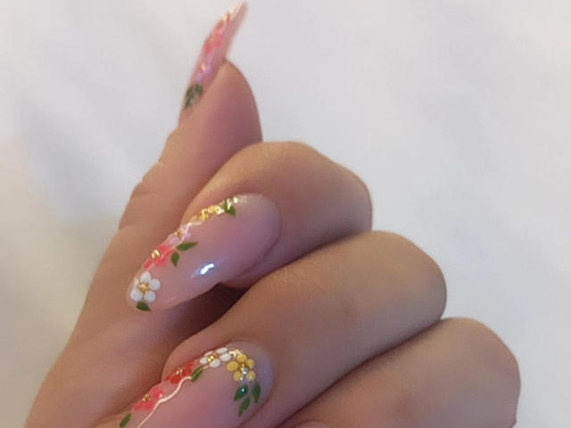 ChristineBray's hand with pink nails, long and sharp, adorned with tiny white flowers, green leaves, and gold glitter, against a plain white background.