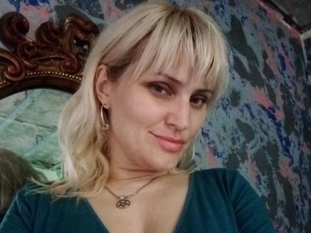 Blonde FreyaMillyy with bangs, green top, small tits, subtle smile, silver necklace, earrings, mirror with wooden frame, textured wall in background.