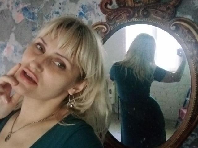 FreyaMillyy, con cabello rubio, una camisa verde y aretes, tocándose el labio, se refleja en un espejo ornamentado que muestra su espalda y trasero. El espejo muestra que ella está sosteniendo un teléfono..