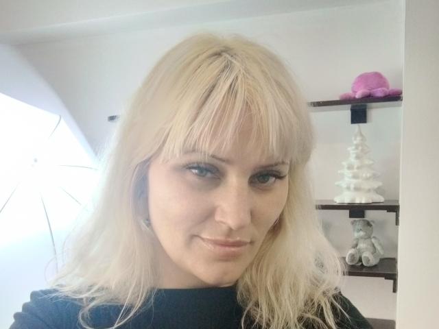 FreyaMillyy, rubia, con flequillo, ojos azules, una ligera sonrisa, una blusa negra, piel clara, un aro nasal discreto, y un pequeño árbol de Navidad blanco y adorable en una estantería al fondo..