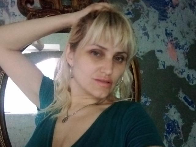 FreyaMillyy con cabello rubio, camisa verde y collar, con la mano en el pelo, mirando a la cámara, busto mediano, labios ligeramente fruncidos, papel tapiz floral azul, espejo en el fondo, maquillaje sutil..