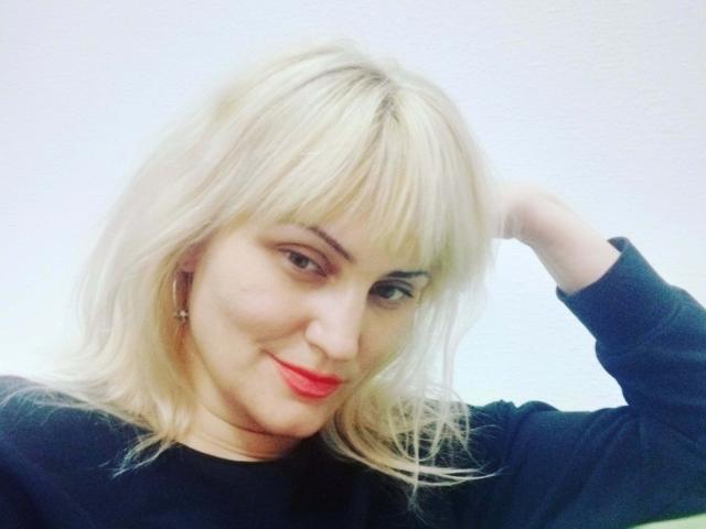 FreyaMillyy a les cheveux blonds, du rouge à lèvres et un pull noir, avec une main dans les cheveux, et elle sourit à la caméra. Ses seins ne sont pas visibles..