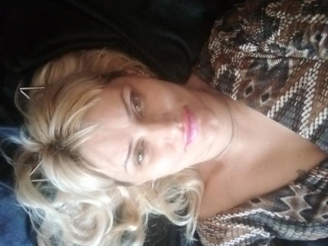 FreyaMillyy hat blondes, welliges Haar, pinken Lippenstift und ein gemustertes braunes Hemd. Sie liegt da, mit einer Metall-Haarspange und einer silbernen Halskette. Ihre Brüste sind teilweise sichtbar..