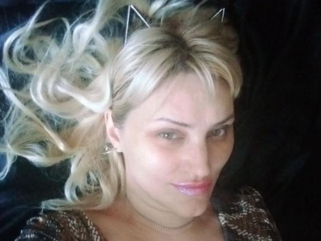 FreyaMillyy, avec ses cheveux blonds, son serre-tête avec des oreilles de chat et ses lèvres roses brillantes, porte un haut marron à motifs et est allongée, ses cheveux ondulés s'étalant autour de sa tête..