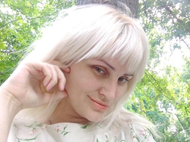 FreyaMillyy met blond haar, een pony en een subtiele glimlach, die haar hand met roze nagels laat zien, gekleed in een bloemenprint top, in een zonnig bos. Haar achterkant is niet zichtbaar..