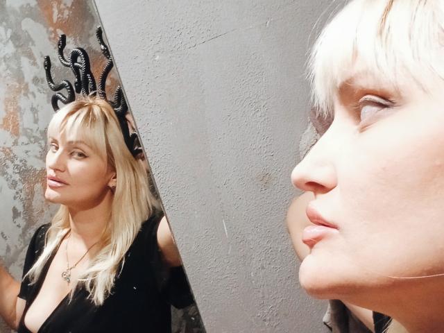 FreyaMillyy con i capelli biondi, indossa una maglietta nera, mostra i suoi seni in uno specchio, il suo riflesso a destra, guarda in alto con le labbra leggermente dischiuse, sfondo di parete con texture..