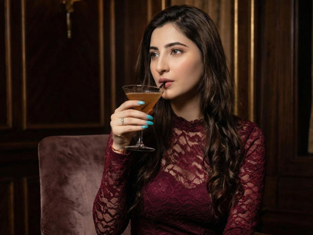 ZoeHillss with long brown hair, lace burgundy top, blue nails, sipping a martini, slight pout, wooden wall background, sitting in a purple velvet chair.