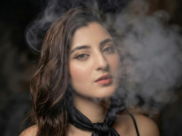 Zoe Hillss, con aliento ahumado, cabello oscuro y ondulado, delineador de ojos sutil y una bufanda negra, tiene una expresión suave y ligeramente seductora, mirando ligeramente hacia un lado..