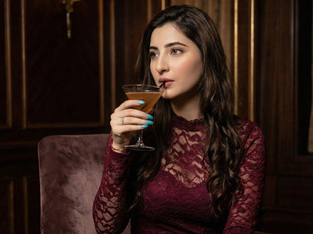 Zoe Hill, con i suoi lunghi capelli castani, sorseggia un cocktail, indossa una blusa color borgogna in pizzo, ha le unghie color turchese e porta un anello, seduta su una poltrona in velluto..