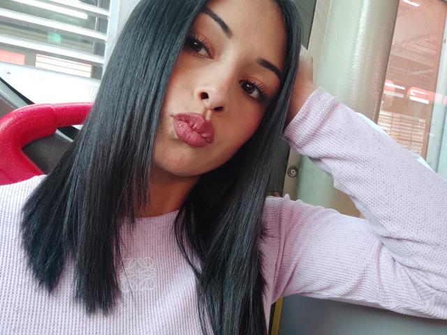 EmilyVv is a young woman with straight, black hair parted in the middle, wearing a light purple, rib-knit sweater. She is making a pouty face with slightly pursed pink lips. Her right hand is touching her hair. She has dark, arched eyebrows and long, thick eyelashes. The light in the image highlights her smooth, medium-brown skin. The composition focuses on her face and upper body. The texture of the sweater contrasts with her smooth hair and skin. The image has a casual, selfie-style quality.