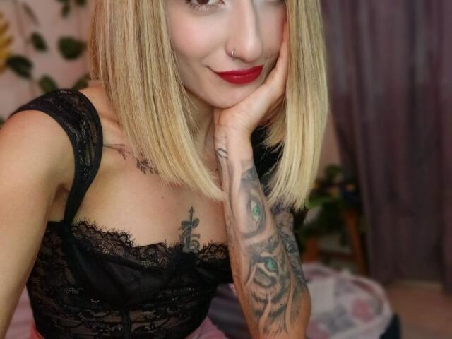 Blonde Kaiix with red lipstick, black lace top, tattooed arm, nose ring, hand on cheek, smiling at camera, showing small tits and a cute face.