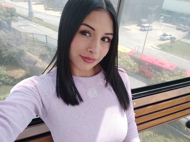 EmilyVv is a young woman with straight black hair, medium length, and a fair complexion. She wears a light purple, rib-knit, long-sleeve top with a small, white embroidered logo on the chest. Her eyebrows are dark and well-defined, and she has long, thick, dark eyelashes. Her lips are full and painted a deep pink. She's taking a selfie from a high angle, looking slightly upward. The background shows a cityscape through a window with a street, red truck, and greenery. The image has a watermark in the bottom right corner.